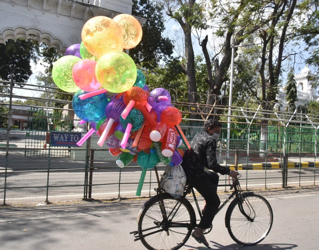 Man wearing mask selling balloons passes assembly road on cycle